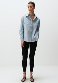 Camicia button-up azzurro chiaro con colletto, maniche lunghe e vestibilità rilassata, abbinata a jeans skinny neri e tacchi a spillo neri.