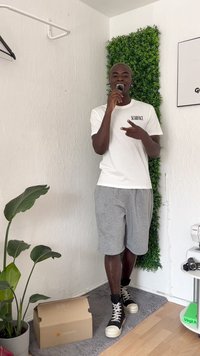 Person, der ein weißes Baumwoll-T-Shirt mit der Aufschrift "SCARFACE", graue Shorts und schwarze High-Top-Sneaker trägt, und neben einer grünen Pflanze sowie einer Box steht.
