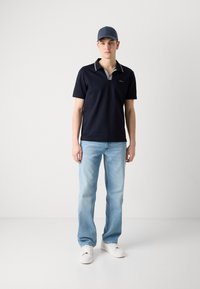 Camisa polo azul marino con cuello contrastante, jeans azul claro y zapatillas blancas. El conjunto presenta un ajuste casual y elementos de diseño simples.