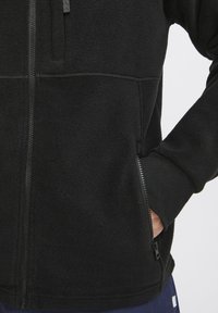 Schwarze Fleecejacke mit Reißverschluss auf der Vorderseite, hohem Kragen, gerippten Bündchen und Reißverschlusstaschen. Glatte Textur mit sichtbaren Nahtdetails.
