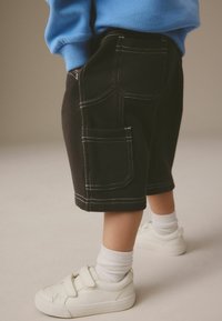 Zwarte katoenen shorts met witte stiksels en zakken, gecombineerd met witte klittenbandschoenen en witte sokken. Blauwe sweatshirt zichtbaar.