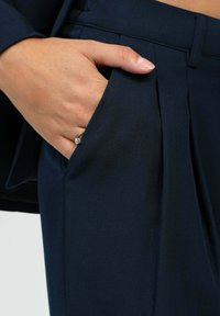 Main avec une bague en argent reposant à l'intérieur de la poche d'un pantalon plissé bleu marine sur un fond clair.