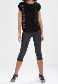 Černé tričko s krátkými rukávy a vertikálním nápisem, kombinované s tmavě šedými capri legínami a šedými sportovními botami. Jednoduchý, přiléhavý design.