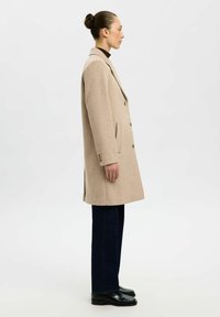Beige wollen jas met een getailleerd ontwerp, zijzakken en een sluiting met drie knopen, gecombineerd met donkere denim broek en zwarte enkellaarsjes.
