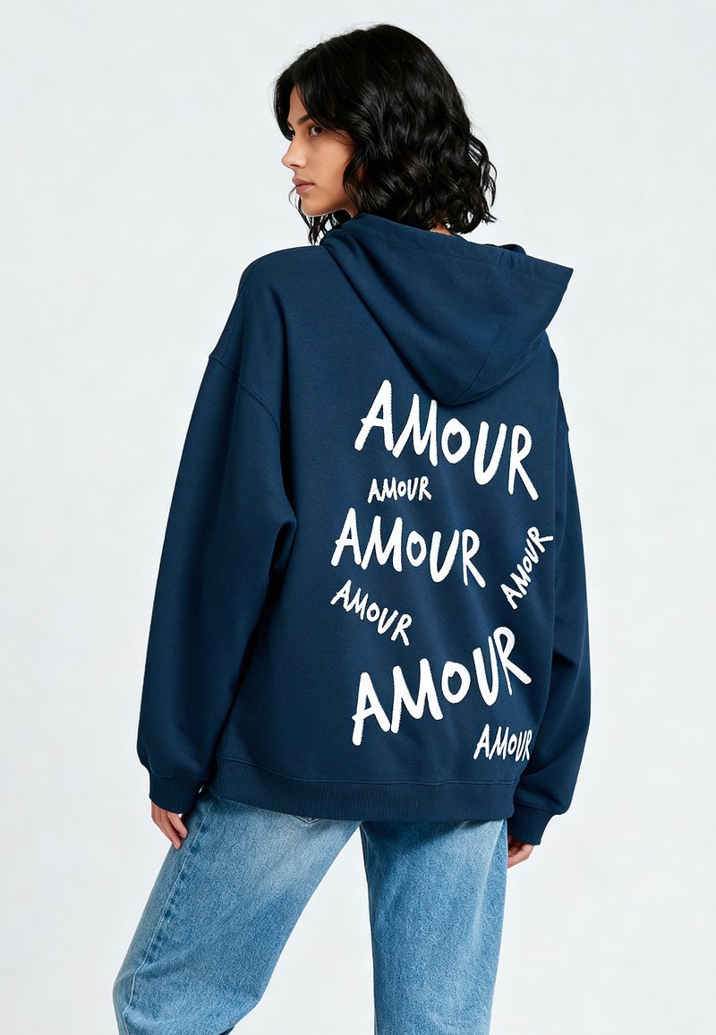 Felpa oversize blu navy con tasca frontale e cappuccio regolabile con coulisse. Testo "AMOUR" bianco di dimensioni variabili stampato sul retro.