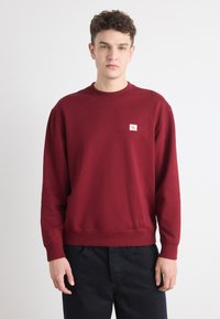 Felpa color bordeaux in cotone con scollatura a girocollo. Presenta polsini e orlo ribattuti, con un piccolo logo bianco ricamato sul petto.