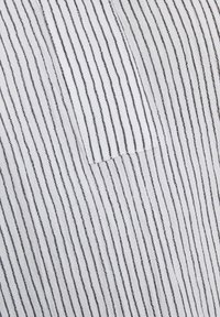 Tissu blanc avec des rayures noires verticales, comportant une petite poche cousue sur le côté. Texture douce, design simple et lignes épurées.