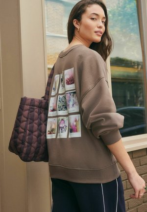 Donna che porta una borsa trapuntata indossa un felpa marrone con stampe fotografiche di fiori sulla schiena, in piedi accanto a un edificio con finestre.