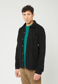Man met kort krullend haar in een zwart knopenjasje over een groen shirt en een bruine broek, staand voor een effen lichte achtergrond.