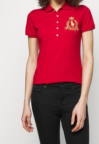 Polo rouge à manches courtes, avec un logo brodé en or et un détail de couronne. Associé à un jean noir ajusté.