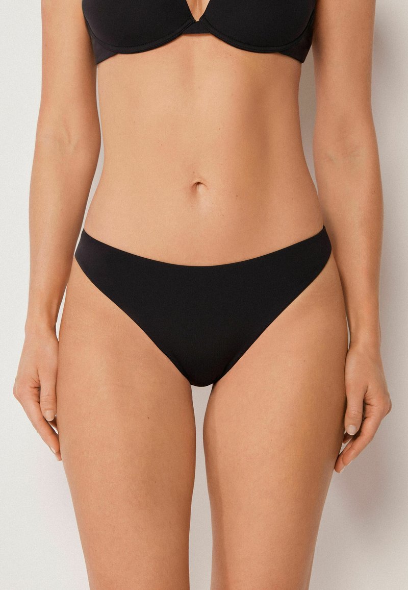 Kvinde iført sort bikini underdel med høj udskæring og matchende BH, stående mod en ensfarvet lys baggrund, forfra.