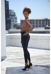 Braune Langarmbluse kombiniert mit schwarzen karierten, figurbetonten Hosen und schwarzen High Heels. Glatter Stoff und maßgeschneiderter Schnitt. Klarer urbaner Hintergrund.