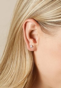 Silberne Steckerohrringe mit drei kleinen klaren Kristallen, die in Blumenform angeordnet sind, getragen am Ohrläppchen eines menschlichen Ohrs mit geradem blonden Haar.