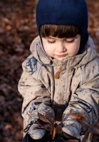Beige jas met patroon en blauwe accenten, bijpassende handschoenen, houdend bruine herfstbladeren tegen een achtergrond van gevallen bladeren.
