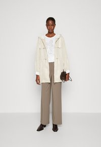 Giacca beige texturizzata con cappuccio e cordoncino, indossata sopra una maglietta bianca con grafiche. Pantaloni marrone a gamba larga e stivali alla caviglia marrone scuro.