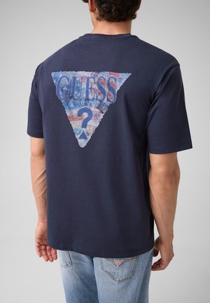 Homme portant un T-shirt bleu marine avec un logo triangle du drapeau américain délavé et le texte "GUESS?" dans le dos, associé à un jean bleu clair.