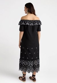 Schwarzes schulterfreies Kleid mit Rüsche am Ausschnitt, das weiße Stickmuster am Saum und geometrische Akzente in der Taille aufweist.