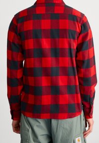 Chemise en flanelle à carreaux rouge et noir avec col boutonné, présentant un tissage texturé et des manches longues avec poignets à boutons.