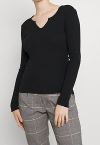 Schwarzes, gerip pt long-sleeve V-Ausschnitt-Oberteil mit strukturierter Oberfläche, kombiniert mit karierten Hosen, die ein helles und dunkles Karomuster aufweisen.