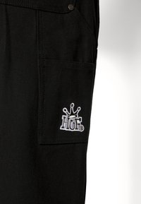 Svarta canvas cargobyxor med en sidficka med en broderad "HUF"-logotyp och krona i vitt. Slät textur och robust design.