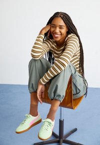 Chemise à manches longues rayée verte et grise, pantalon ample olive, baskets vert clair avec des accents roses, assis sur une chaise en bois devant un fond uni.