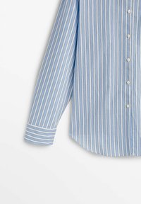 Chemise bleu clair à manches longues avec rayures blanches verticales, boutonnage sur le devant et ourlet arrondi. Le poignet présente un détail rayé contrastant.