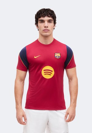 Muž v červeno-tmavomodrom drese FC Barcelona so žltým logom Spotify a bielymi šortkami stojaci pred jednoliatym pozadím.