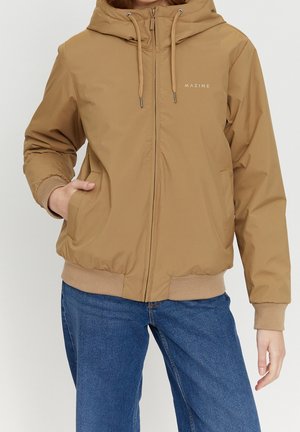 Personne portant une veste à capuche beige avec une fermeture éclair à l'avant et des poches, associée à un jean bleu, debout avec une main dans une poche.