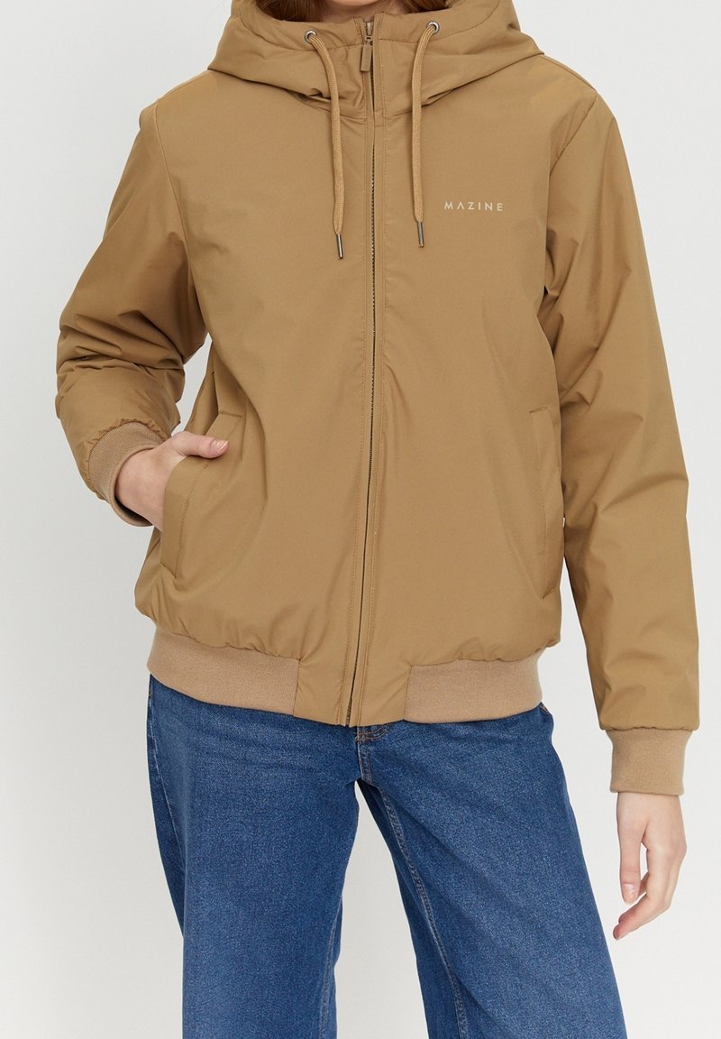 Personne portant une veste à capuche beige avec une fermeture éclair à l'avant et des poches, associée à un jean bleu, debout avec une main dans une poche.