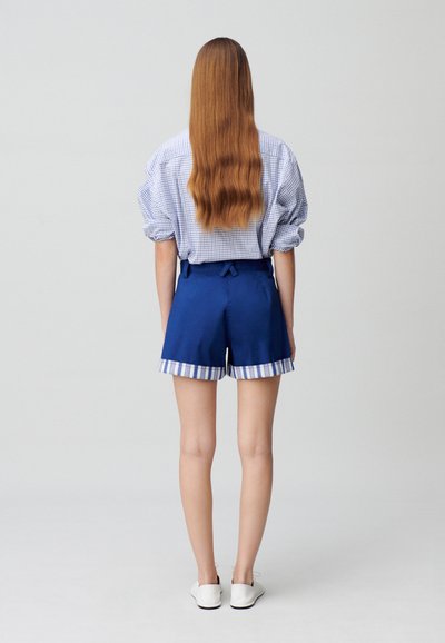 Shorts bleus avec un ourlet rayé et un détail en nœud à la taille, associés à une chemise à carreaux. Tissu lisse, design décontracté.