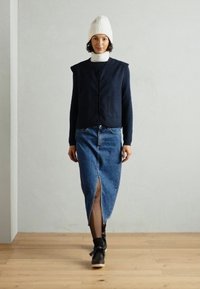 Marineblauwe streepvesten, witte coltrui, denimrok met voorknoop en zwarte schoenen. Model staat op een houten vloer tegen een lichte muur.