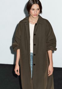 Cappotto lungo e largo di colore verde oliva, realizzato in tessuto leggero, con colletto, bottoni frontali e tasche laterali.