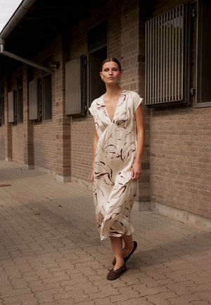 Nő sétál egy burkolt úton egy téglából épült épület mellett, bézs ruhát visel barna levélmintával és sötétbarna bebújós cipőket.