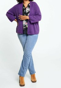 Cardigan viola lavorato a maglia con vestibilità ampia, indossato sopra una camicetta nera a fiori. Abbinato a jeans di denim chiaro e stivaletti marrone chiaro.