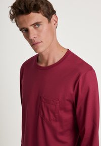 T-shirt bordeaux à manches longues en tissu doux, avec un col rond et une petite poche sur la poitrine gauche. Design simple, texture lisse.