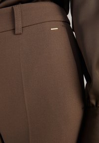 Gros plan sur un pantalon marron texturé avec une boucle de ceinture et un petit détail rectangulaire doré près de la taille, porté avec un haut marron assorti.