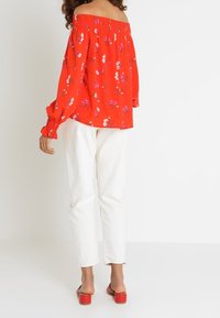 Červená off-the-shoulder blůza s květinovým vzorem, dlouhé rukávy s elastickými manžetami, kombinovaná s bílými kalhotami a červenými sandály na vysokém podpatku.
