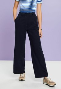 Marinblå vida byxor med framfickor, bälteshällor och en slät yta, i kombination med en randig blå och vit topp samt beiga sneakers.