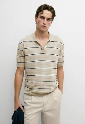 Giovane uomo con i capelli mossi che indossa una polo beige a righe con maniche corte e pantaloni chiari, tenendo una giacca scura in una mano.