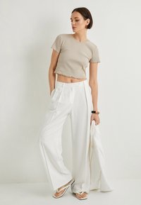Top corto de canalé beige condo un dobladillo en forma de ondulación, combinado con pantalones de pierna ancha blancos de cintura alta. La modelo sostiene una chaqueta blanca y lleva chanclas.