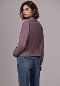 Ljuslila bomberjacka med en dragkedjeficka på vänster ärm och elastiska muddar. Buren över blå denim jeans med en avslappnad passform.