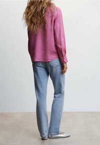 Femme aux longs cheveux ondulés portant un chemisier rose à manches longues, un jean bleu clair et des ballerines argentées, debout devant un fond uni.