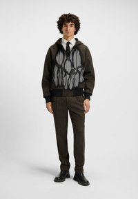 Brązowy hoodie na zamek z szarym abstrakcyjnym wzorem z przodu, zestawiony z teksturowanymi brązowymi spodniami i czarnymi botkami za kostkę. Model stoi wyprostowany.