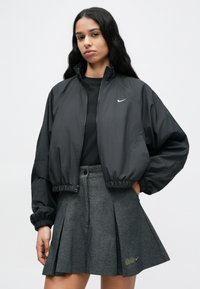 Μαύρο bomber μπουφάν με ελαστικά μανίκια, με μικρό λευκό λογότυπο Nike. Συνδυασμένο με γκρι πλισέ φούστα, που δείχνει υφή.