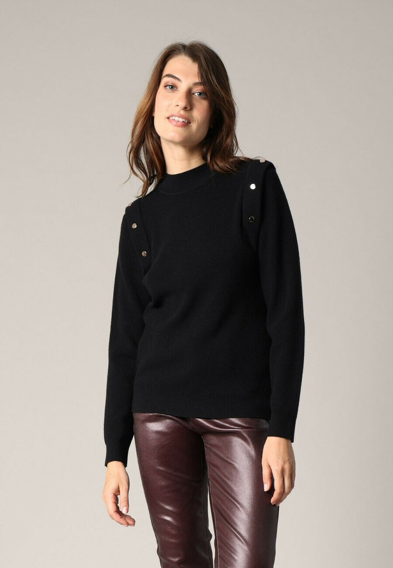 Vrouw in een zwarte trui met zilveren knopen op de schouders en maroon leren broek, staande tegen een effen beige achtergrond.