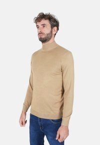 Maglione a collo alto beige in morbido materiale a maglia, design aderente, maniche lunghe, polsini e orlo a coste, abbinato a jeans scuri.