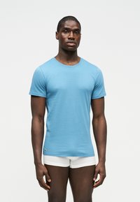 Άνδρας που φοράει εφαρμοστό ανοιχτό μπλε κοντομάνικο μπλουζάκι και λευκά μποξεράκια, στέκεται μπροστά σε απλό λευκό φόντο.