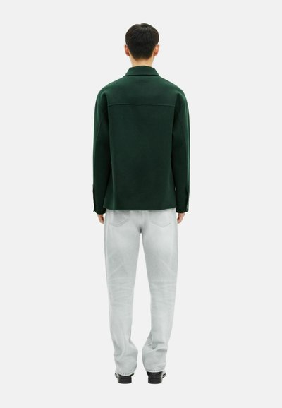 Man staat met zijn gezicht naar de andere kant, gekleed in een donkergroen langemouwenshirt, lichtgrijze spijkerbroek en zwarte schoenen, tegen een witte achtergrond.