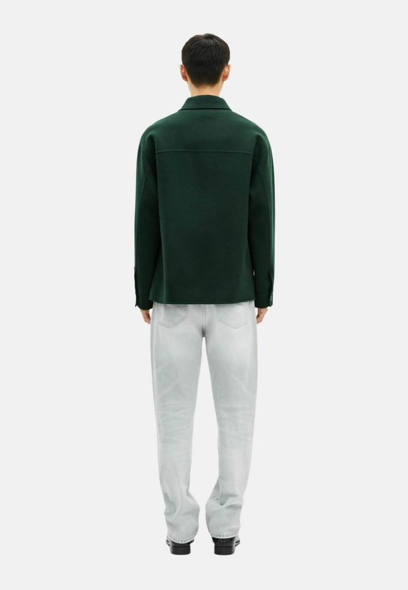 Mand stående med ryggen til iført en mørkegrøn langærmet skjorte, lys grå jeans og sorte sko på en hvid baggrund.