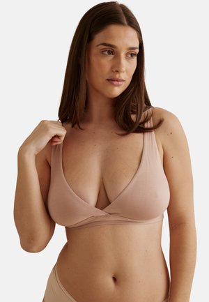 Femme aux longs cheveux bruns portant un soutien-gorge sans manches beige clair et une lingerie assortie, regardant légèrement vers la droite.